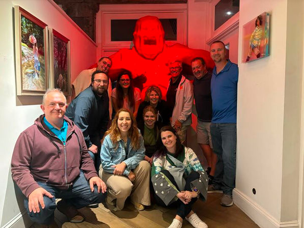 Group of a dozen adults smiling in an indoor hallway/gallery posing for a playful photo-op in front of a large red monster figure with framed artwork on the walls.