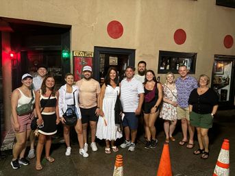 Smiling group of adults in casual summer clothes posed outside a lit tavern entrance on a wet city sidewalk at night, tan stucco wall with red circular accents and orange traffic cones in the foreground.