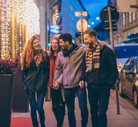 Four friends laughing as they walk arm-in-arm along a downtown city sidewalk at night, wearing winter coats and scarves under festive holiday string lights and storefront decorations