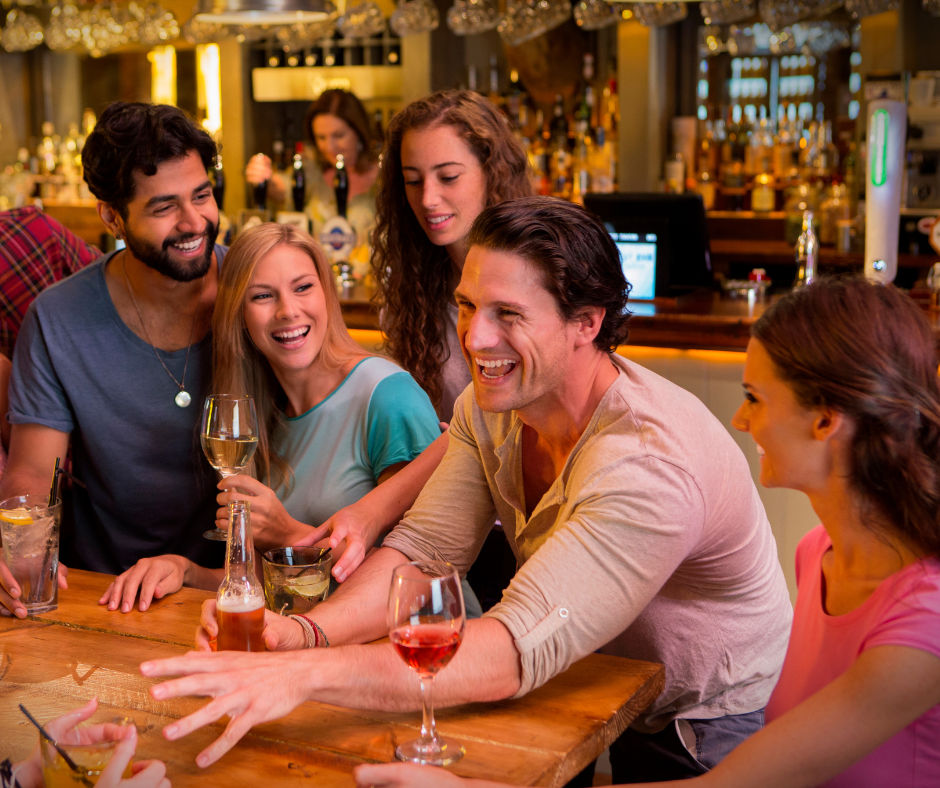 Group of friends laughing and sharing drinks at a lively bar—wooden countertop with wine glasses, beer and cocktails on a fun night out