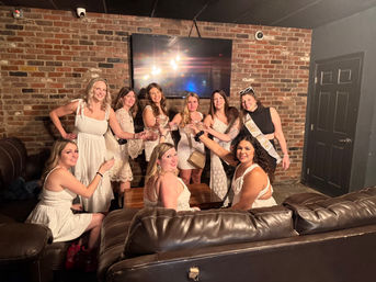 Group of women in white dresses toasting in a cozy brick-walled lounge with leather sofas and a wall-mounted TV — bachelorette party vibe with a "Bride to Be" sash.