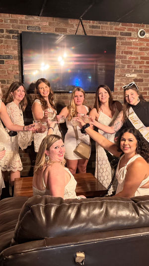 Group of women in white dresses toasting in a cozy brick-walled lounge with leather sofas and a wall-mounted TV — bachelorette party vibe with a "Bride to Be" sash.