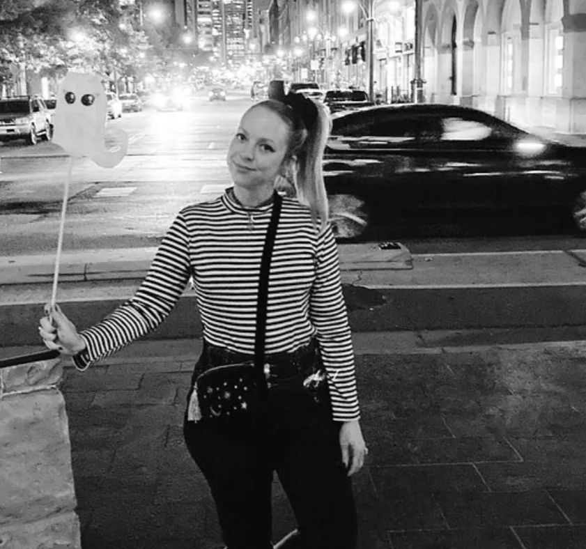 Person in a striped long-sleeve shirt holding a ghost-shaped balloon on a stick, posing on a downtown city sidewalk at night with blurred car traffic and lit buildings in a black-and-white photo.