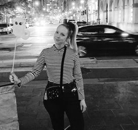 Person in a striped long-sleeve shirt holding a ghost-shaped balloon on a stick, posing on a downtown city sidewalk at night with blurred car traffic and lit buildings in a black-and-white photo.