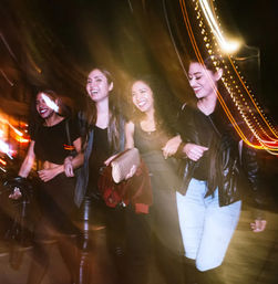 Four women laughing and walking arm-in-arm on a city street at night, motion blur and streaking lights capturing lively urban nightlife.