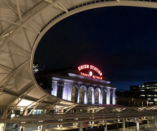 Nighttime view of a historic train station facade lit with purple uplighting and a glowing red neon sign, framed by a sweeping modern canopy at an urban rail hub.
