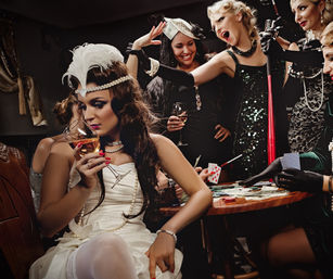 Roaring Twenties–style flapper women in glamorous dresses and pearls sipping cocktails and gambling with cards, chips and cash at a vintage speakeasy table.