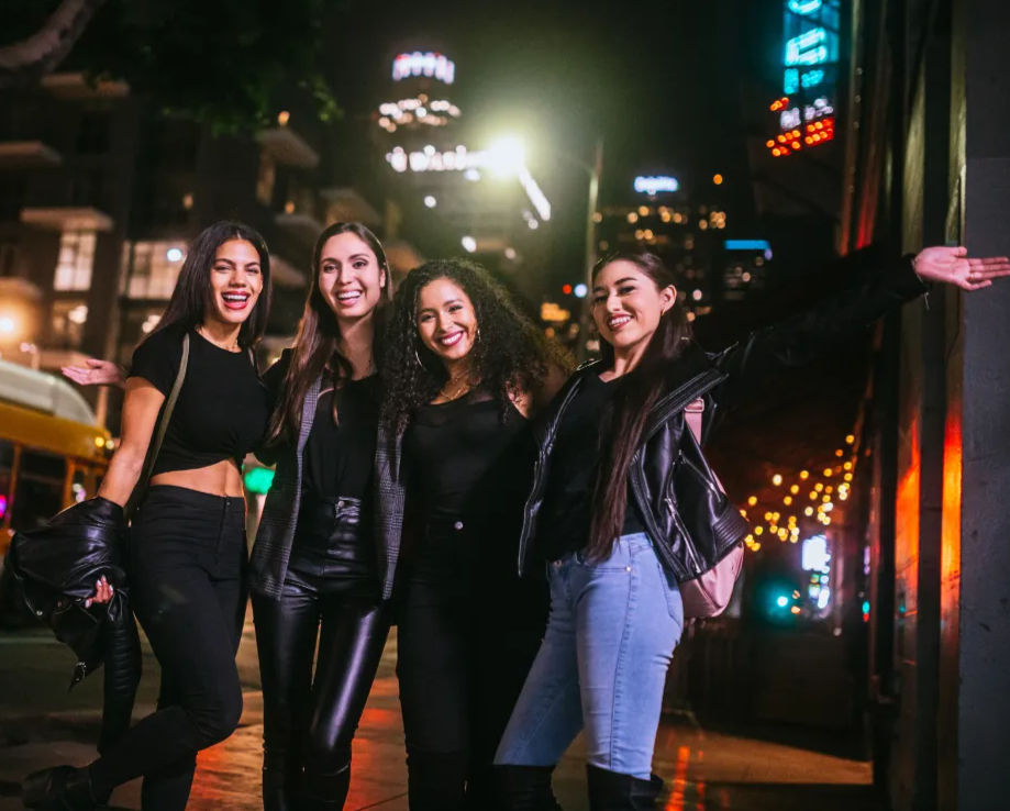 Four friends smiling and posing on a downtown city street at night under neon signs and string lights, enjoying a fun urban night out.
