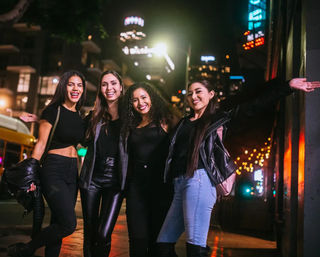 Four friends smiling and posing on a downtown city street at night under neon signs and string lights, enjoying a fun urban night out.