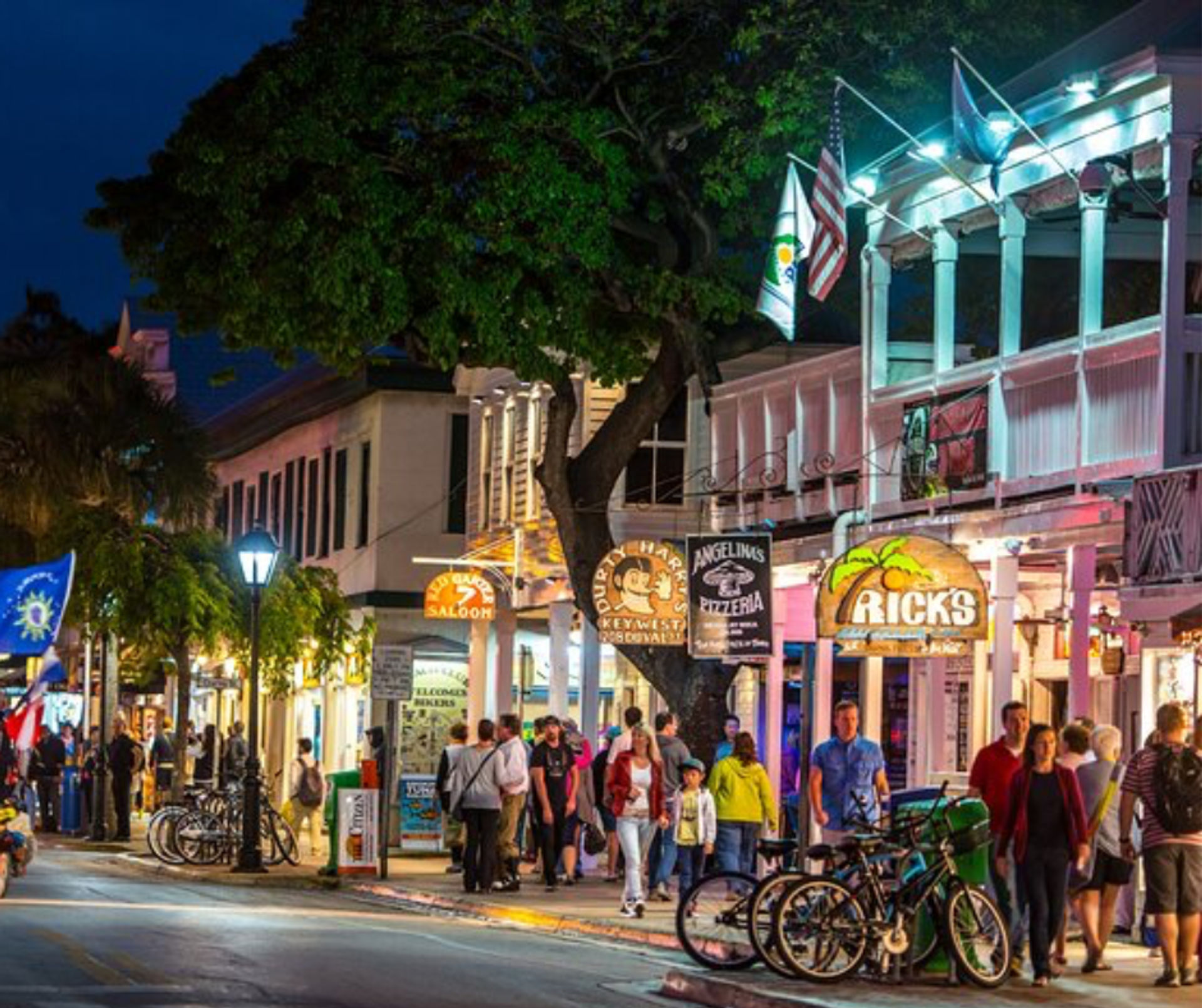 Lively tropical Key West, Florida nighttime street scene with illuminated wooden storefronts, hanging flags and signs, palm trees, parked bicycles and crowds of pedestrians strolling the sidewalk.