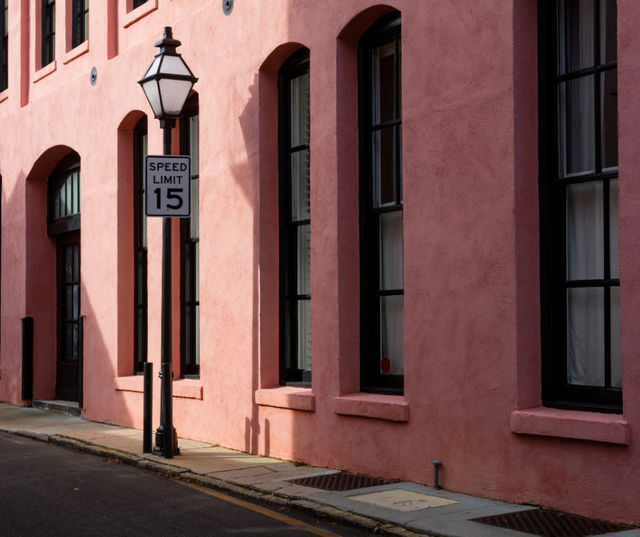 Pink stucco row building with tall arched windows and black trim, a gaslamp-style street lantern and a 15 mph speed limit sign along a narrow city sidewalk.