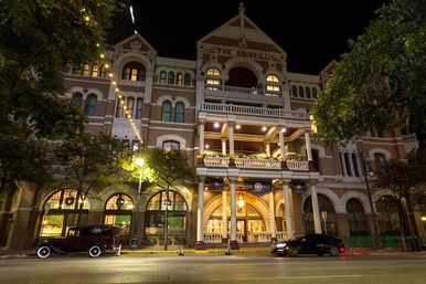 Nighttime downtown scene showing a historic Victorian-style hotel facade with arched windows, ornate balconies, warm exterior lights, string lights and parked cars