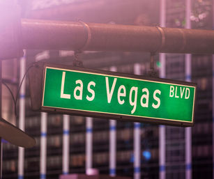 Green Las Vegas Blvd street sign hanging from a metal pole over a neon-lit city street at dusk, with blurred high-rise lights in the background