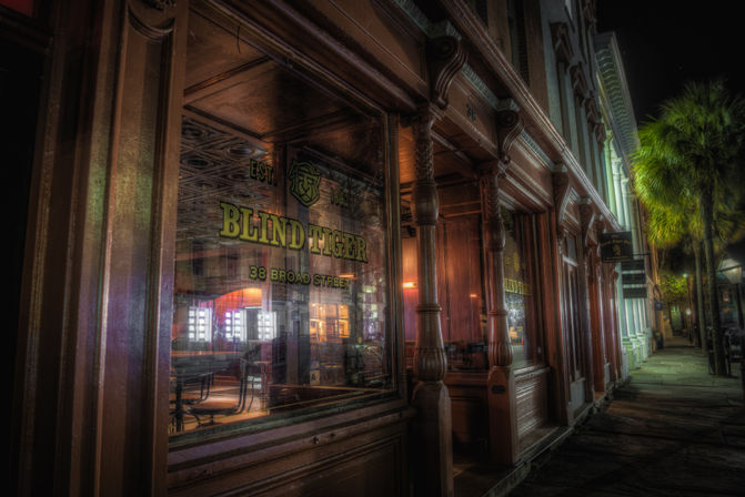 Historic wooden storefront at night with ornate carved columns and large glass windows revealing a warmly lit bar interior, empty tables, and a palm-lined downtown sidewalk.