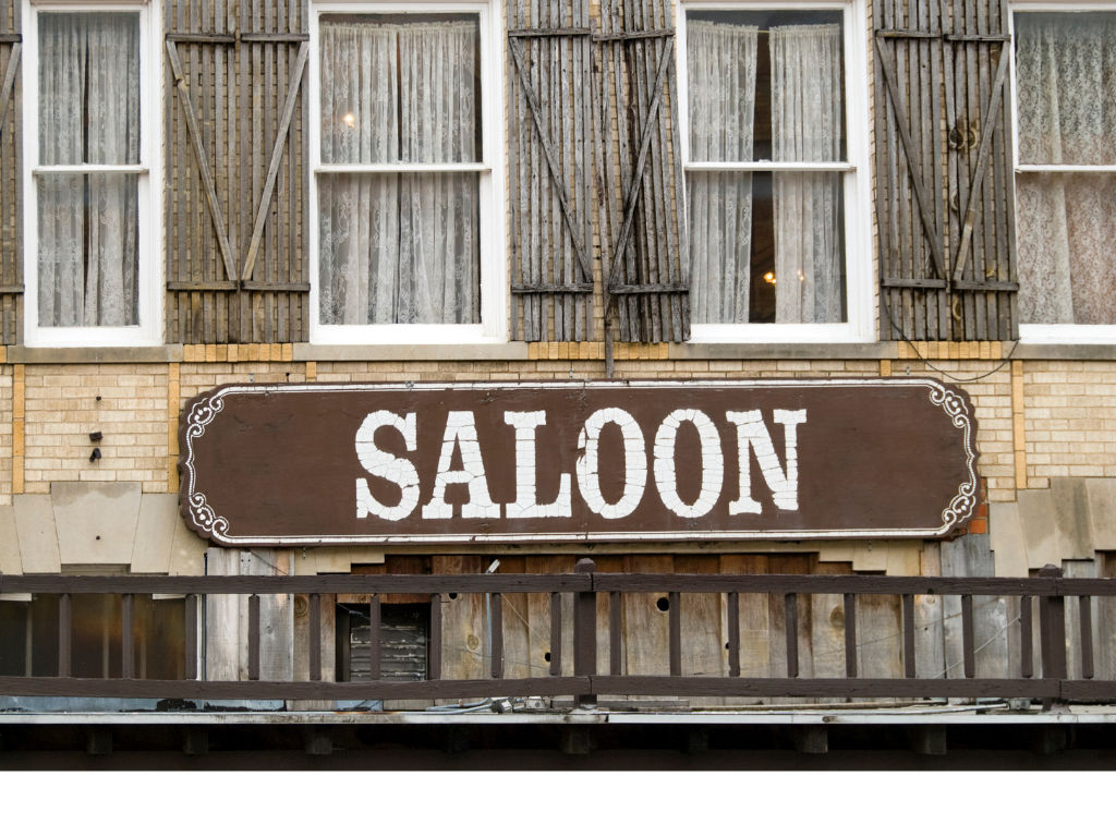 Old-west style wooden 'SALOON' sign on a vintage brick building exterior with lace-curtained windows, weathered shutters, and a worn wooden balcony.