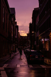 Moody downtown city street at purple sunset — wet pavement reflecting streetlights, brick buildings flanking parked cars, lone pedestrian with an umbrella walking down the center.