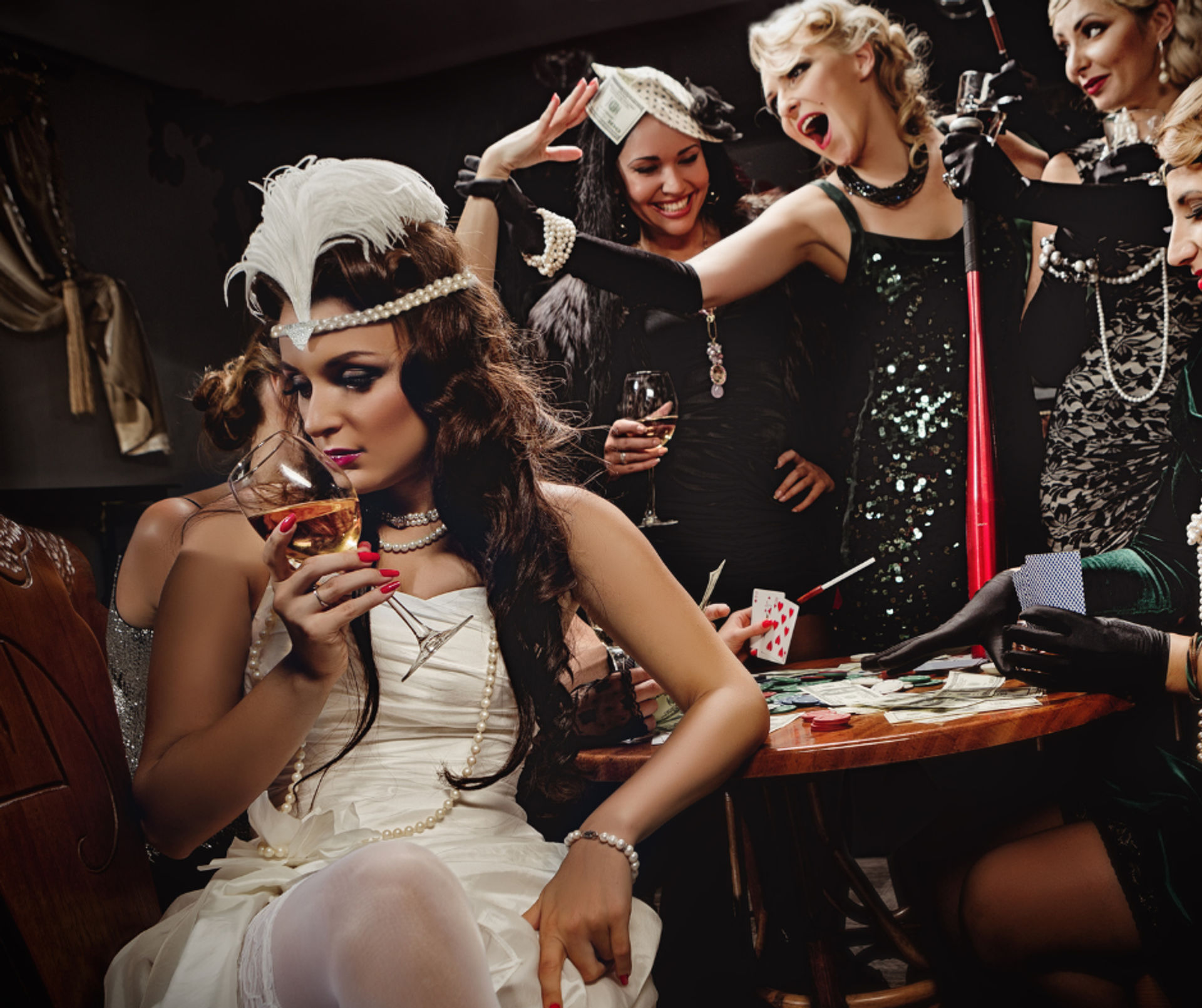 1920s‑style flapper party scene — woman in white feather headband sips wine while friends in sequined dresses gamble, throw cash and play cards at a speakeasy table