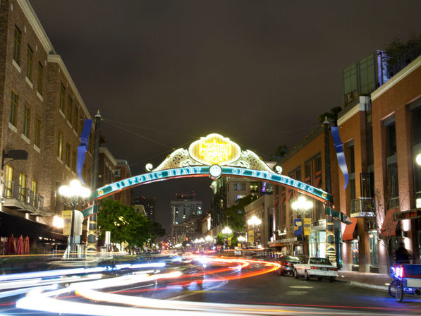 Nighttime scene beneath the glowing Gaslamp Quarter arch in downtown San Diego, with colorful car light trails and historic brick buildings lining the street.