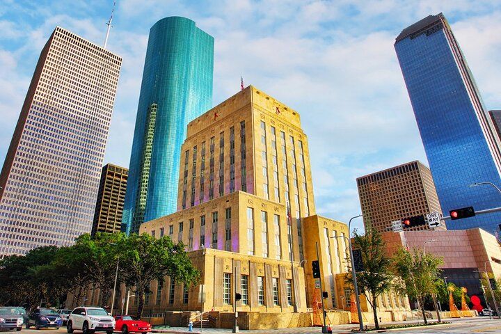 Sunlit Art Deco municipal building framed by a turquoise cylindrical tower and reflective skyscrapers in a downtown skyline, with cars and trees along the city street under a blue sky.