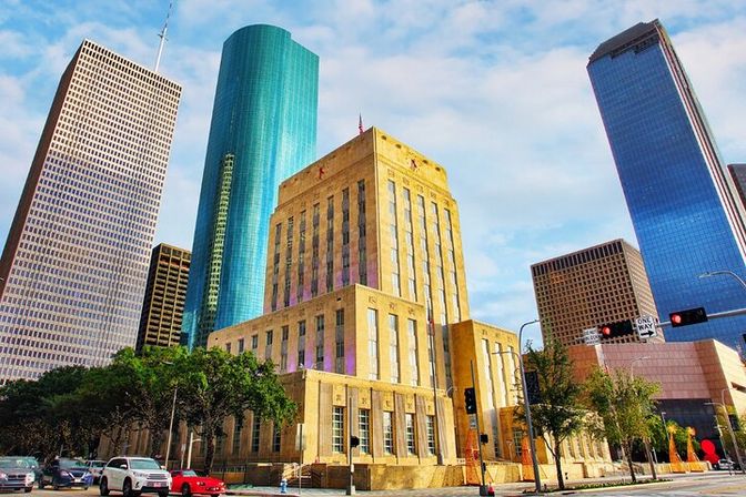 Sunlit Art Deco municipal building framed by a turquoise cylindrical tower and reflective skyscrapers in a downtown skyline, with cars and trees along the city street under a blue sky.