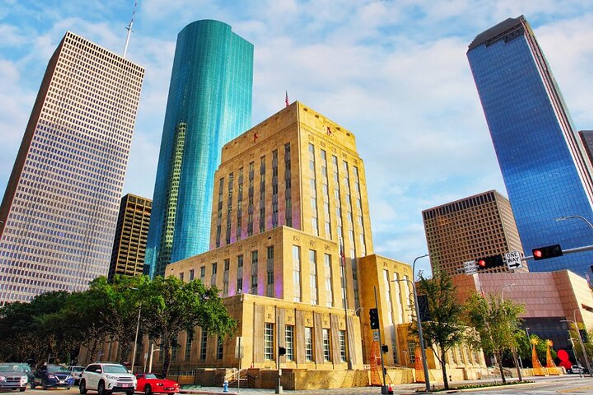 Sunlit Art Deco municipal building framed by a turquoise cylindrical tower and reflective skyscrapers in a downtown skyline, with cars and trees along the city street under a blue sky.