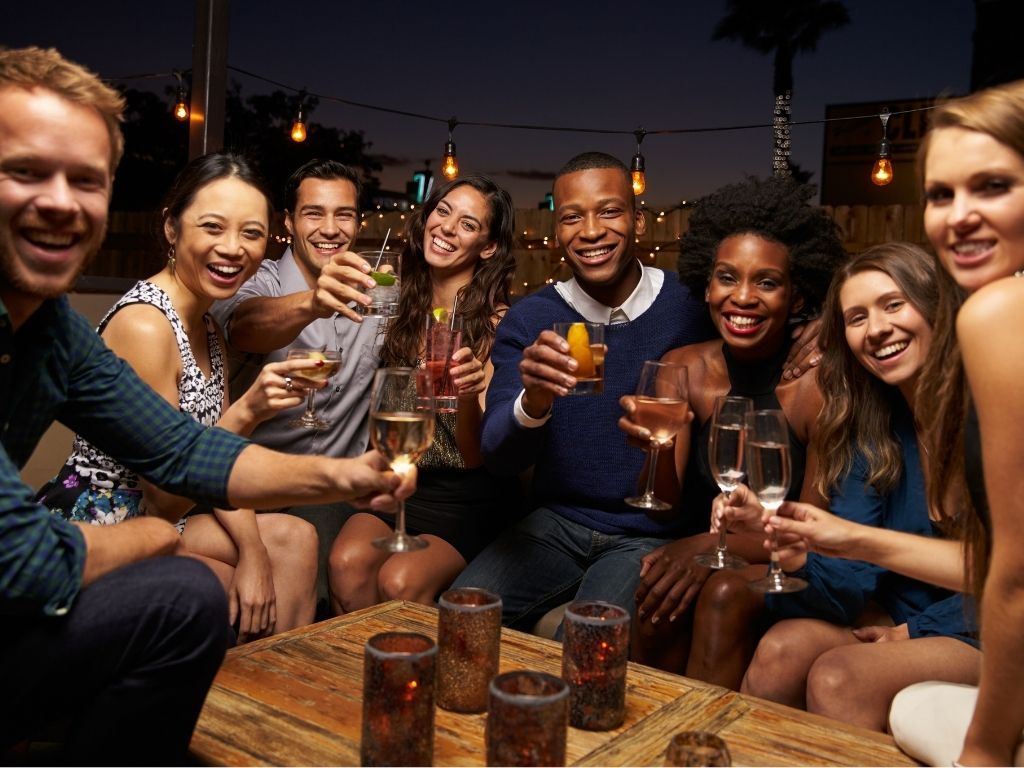 Group of friends laughing and toasting with cocktails and wine on an outdoor patio at night, gathered around a wooden table with candle votives and hanging string lights.