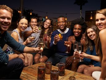 Group of friends laughing and toasting with cocktails and wine on an outdoor patio at night, gathered around a wooden table with candle votives and hanging string lights.