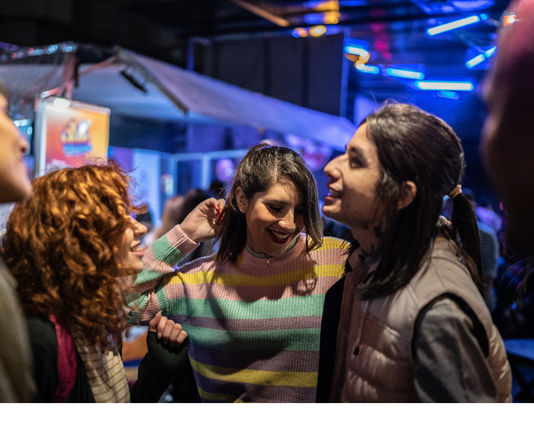 Three friends laughing and dancing under colorful neon lights in a lively indoor bar, urban nightlife scene
