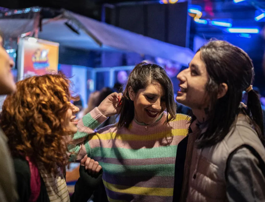 Three friends laughing and dancing during a lively urban night out at an outdoor bar with neon lights — woman in pastel striped sweater center, curly-haired friend left, long-haired friend right.