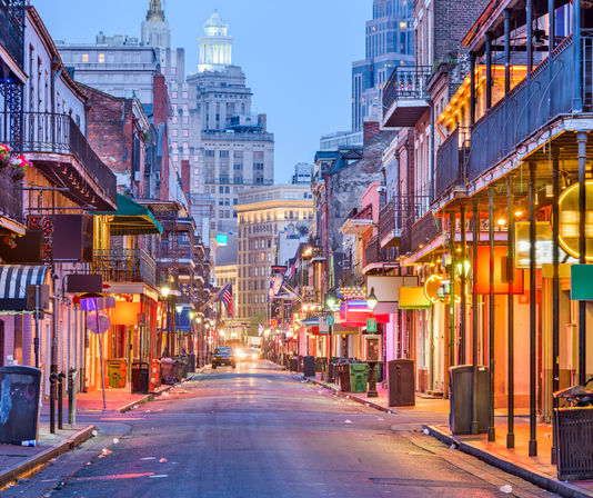 Vibrant Bourbon Street in New Orleans' French Quarter at dusk — empty street lined with wrought-iron balconies, colorful neon signs, and a glowing downtown skyline