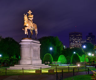 Gilded equestrian statue on a stone plinth lit at night in an urban park with manicured hedges and illuminated city skyline in the background