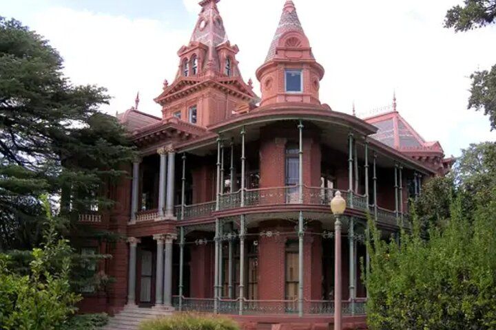 Historic red-brick Victorian mansion with conical turrets, two-story wraparound porch and ornate ironwork, framed by trees and a vintage lamp post.