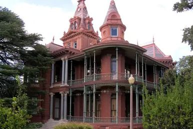 Historic red-brick Victorian mansion with conical turrets, two-story wraparound porch and ornate ironwork, framed by trees and a vintage lamp post.