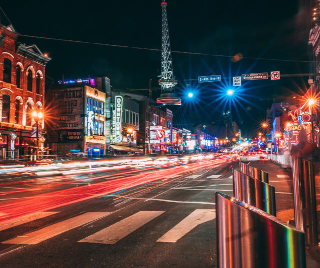 Vibrant downtown nightlife street at night with neon signs on brick buildings, an illuminated spire, blurred car light trails and a crosswalk in the foreground.