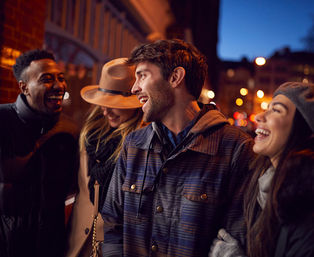 Group of four friends laughing on a city sidewalk at night, bundled in coats and hats with warm streetlights and bokeh city lights in the background — a lively urban night out.