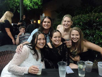 Five smiling women posing together on an outdoor nighttime patio, seated at a table with plastic cups and cocktails, greenery and other patrons visible in the background.