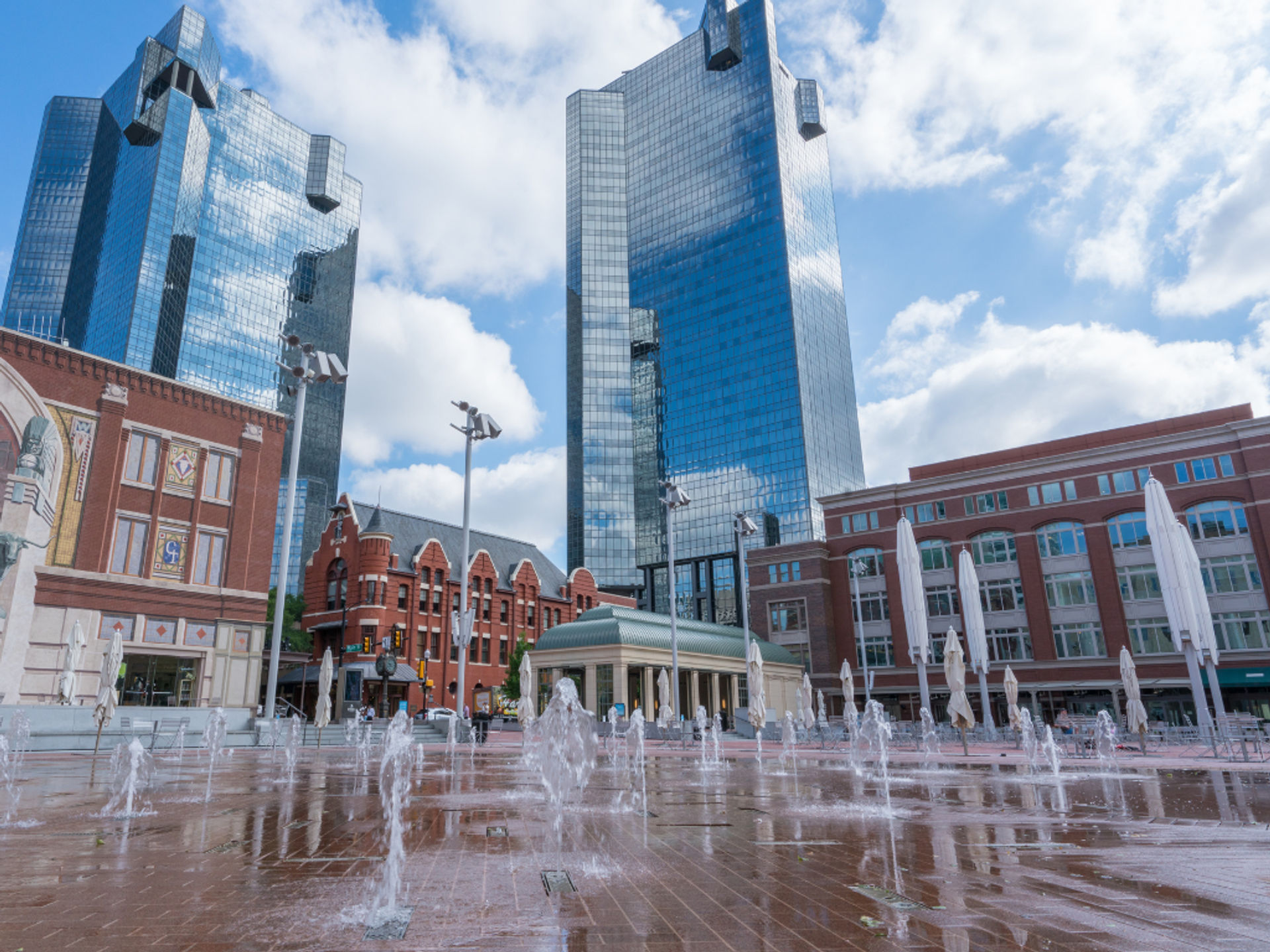 Downtown urban plaza with ground-level splash fountains, reflecting modern glass skyscrapers, historic red-brick buildings and closed patio umbrellas under a bright blue sky.