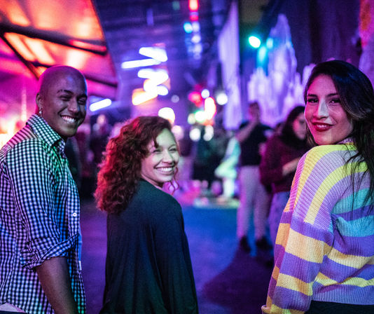 Three friends turn and smile at the camera in a neon-lit downtown nightlife venue, colorful lights and a blurred crowd in the background.