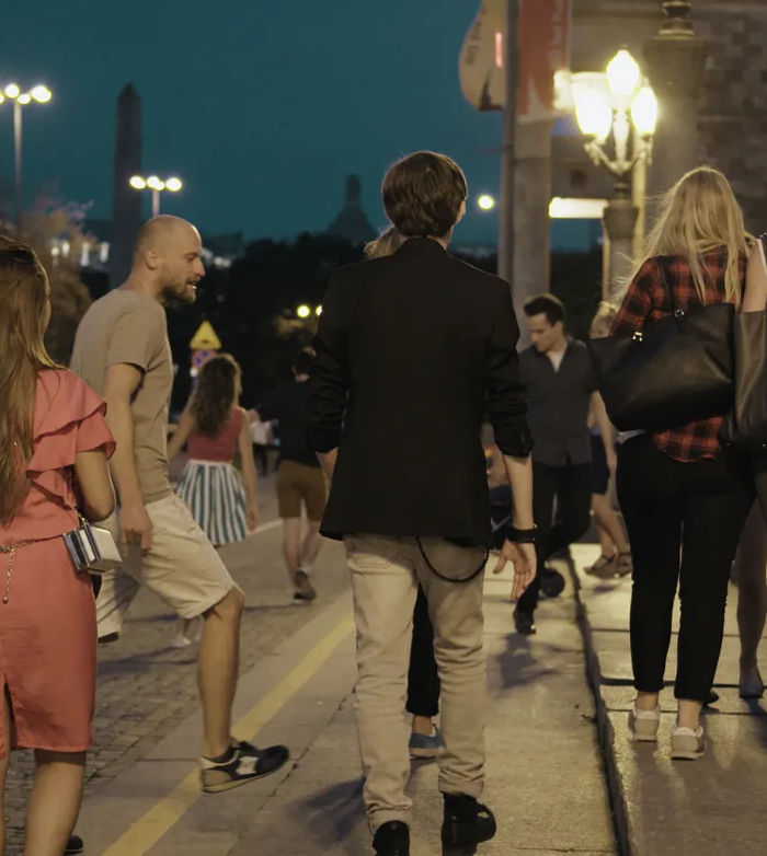 Evening downtown street scene: people in casual summer clothes walking along a cobblestone sidewalk under warm lamp-lit streetlights, urban nightlife vibe