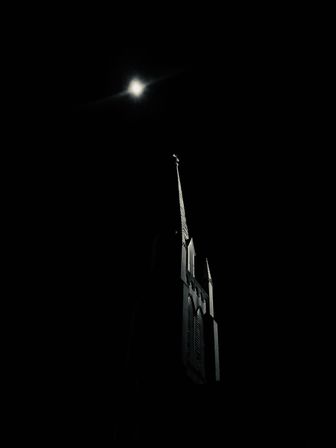 Dramatic moonlit church steeple silhouette with a glowing moon in a pitch-black night sky, Gothic spire and cross faintly illuminated.