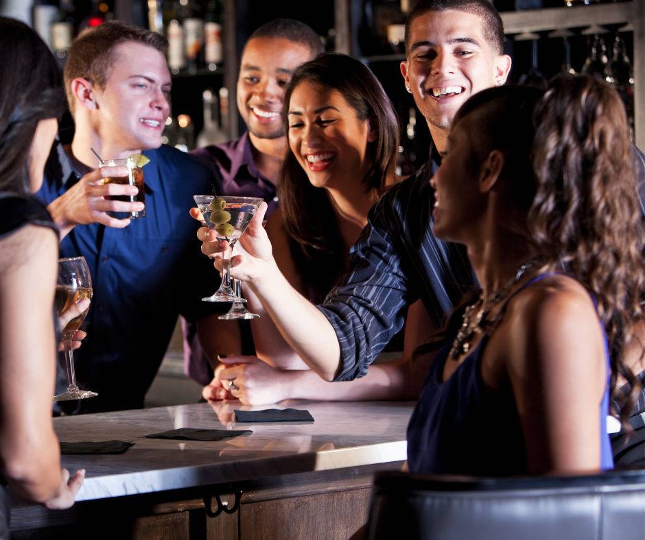 Group of friends laughing and clinking cocktails at a lively bar — martini with olives and wine glasses in upbeat city nightlife.