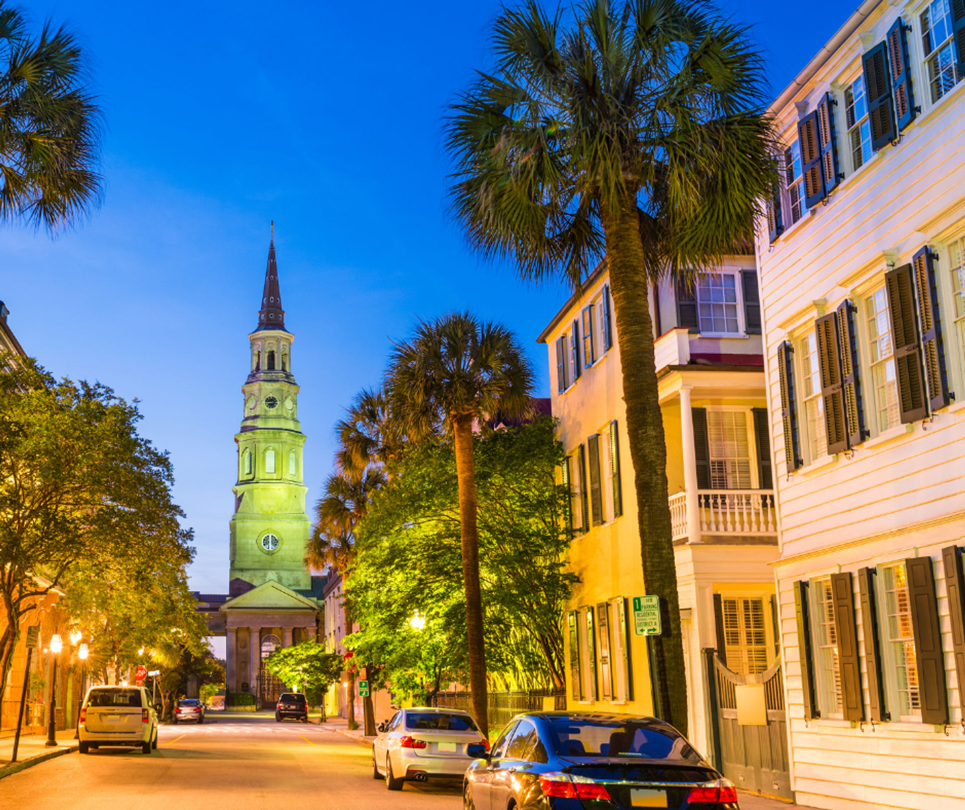 Historic Charleston street at dusk with a glowing church steeple, palm trees, and pastel colonial houses lining a quiet, car-lined road
