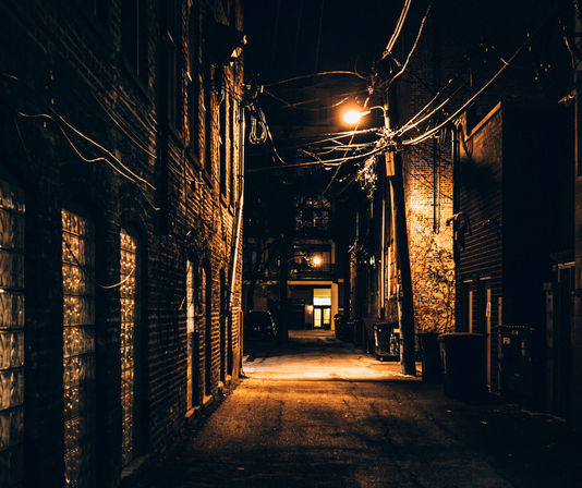 Moody city alley at night—brick walls, tangled overhead utility wires and a lone amber streetlight casting warm light over dumpsters and a distant lit doorway.