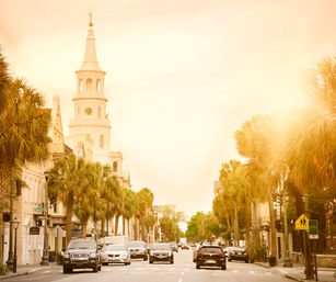 Golden-hour southern coastal downtown street lined with palm trees, historic white church steeple and cars driving along a busy avenue.