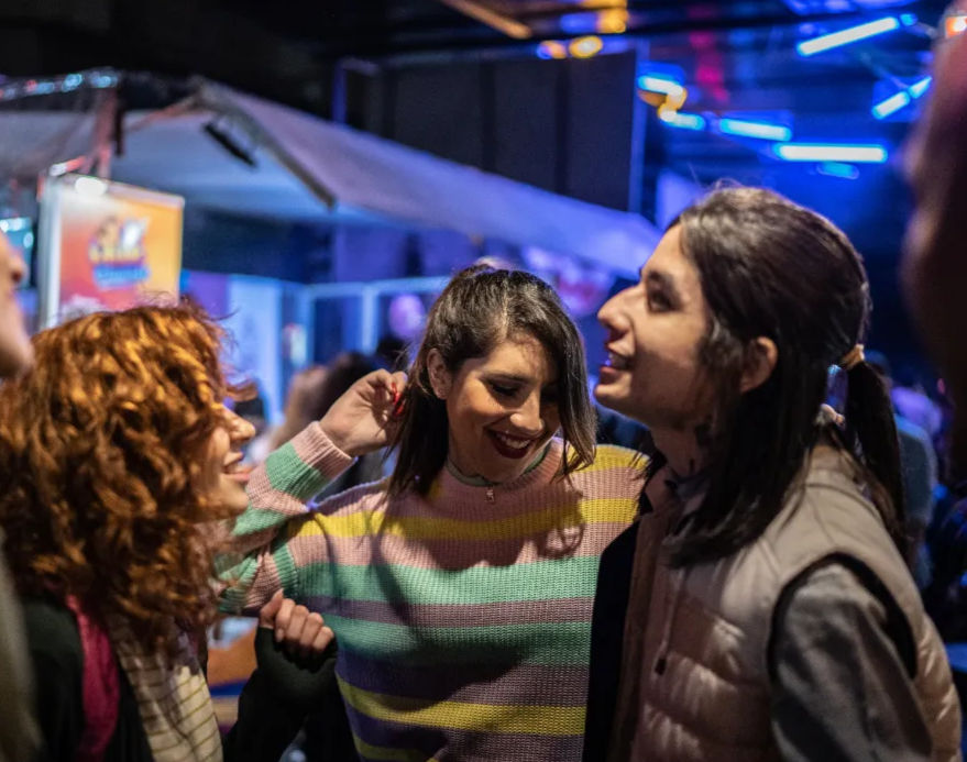 Three friends laughing and dancing under neon lights at a lively indoor nightlife venue, center person wearing a pastel striped sweater.