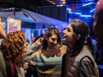 Three young adults laughing and dancing on a night out in a crowded urban bar with colorful neon lights and a lively nightlife vibe.