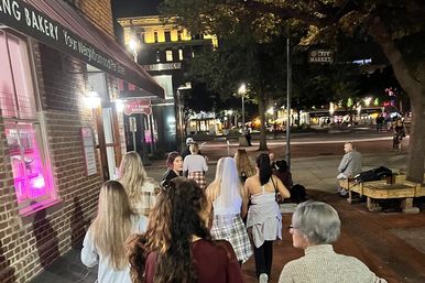 Group of friends walking at night in a downtown plaza past a lit brick bakery storefront; one woman wearing a bridal veil for a bachelorette outing, with trees, benches, and illuminated streetscape in the background.