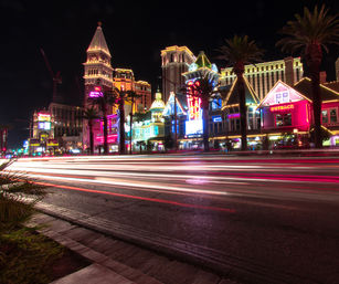 Nighttime Las Vegas Strip scene with colorful neon-lit casino facades, palm trees, and long-exposure streaks of car lights on the boulevard.