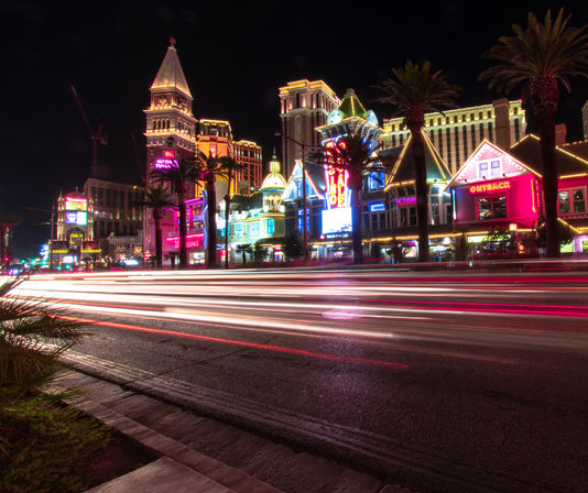 Las Vegas Strip at night — neon-lit casino facades and palm trees with long-exposure light trails streaking across a busy boulevard