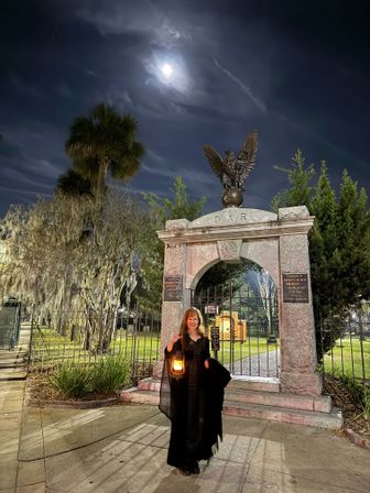 Person in a black cloak holding a lantern beside a stone cemetery arch topped by an eagle statue, moonlit sky and Spanish-moss draped oaks creating a spooky historic Southern cemetery scene at night.